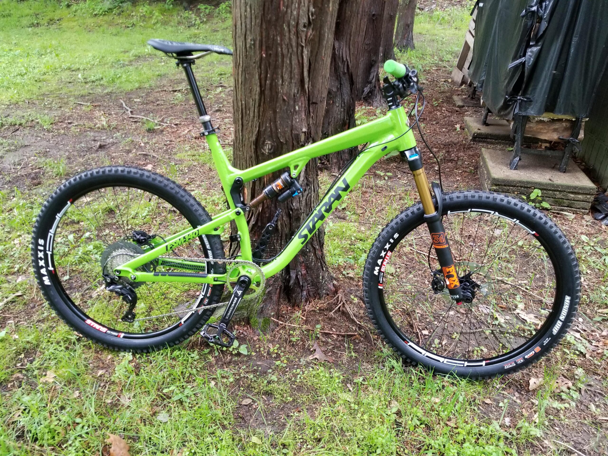 Staran Cycles FSM-140: A bright green mountain bike leaning against a tree in a grassy area, showcasing its features like thick tires, a rear shock, and a prominent fork. The background includes blurred vegetation and a partially visible structure.