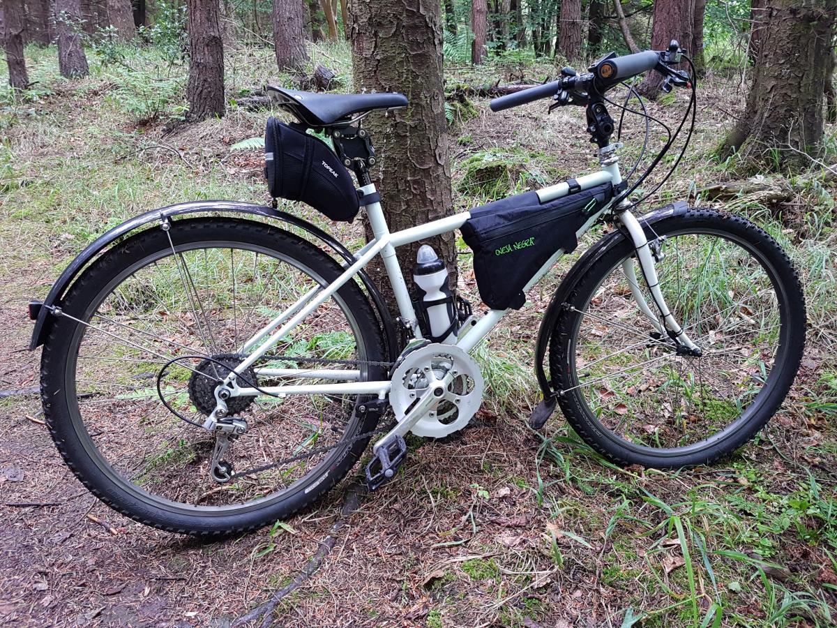 Rocky Mountain Blizzard: A white mountain bike parked on a dirt path surrounded by trees and greenery, featuring a black saddle bag and a water bottle holder. The bike has chunky tires suitable for rough terrain and is positioned against a tree in a forested area.