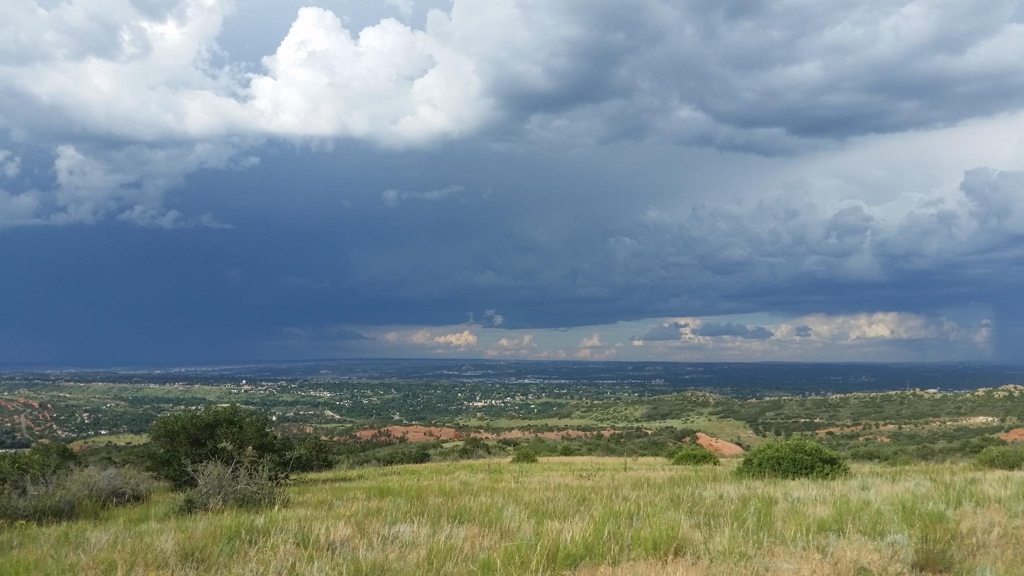 A panoramic view of a landscape under a dramatic sky, featuring dark clouds and hints of blue sky peeking through. The foreground showcases green grass and shrubs, while rolling hills and distant patches of trees create a vibrant backdrop, suggesting an approaching storm with varied shades of gray and blue in the clouds. Red Rock Canyon mountain bike trail.