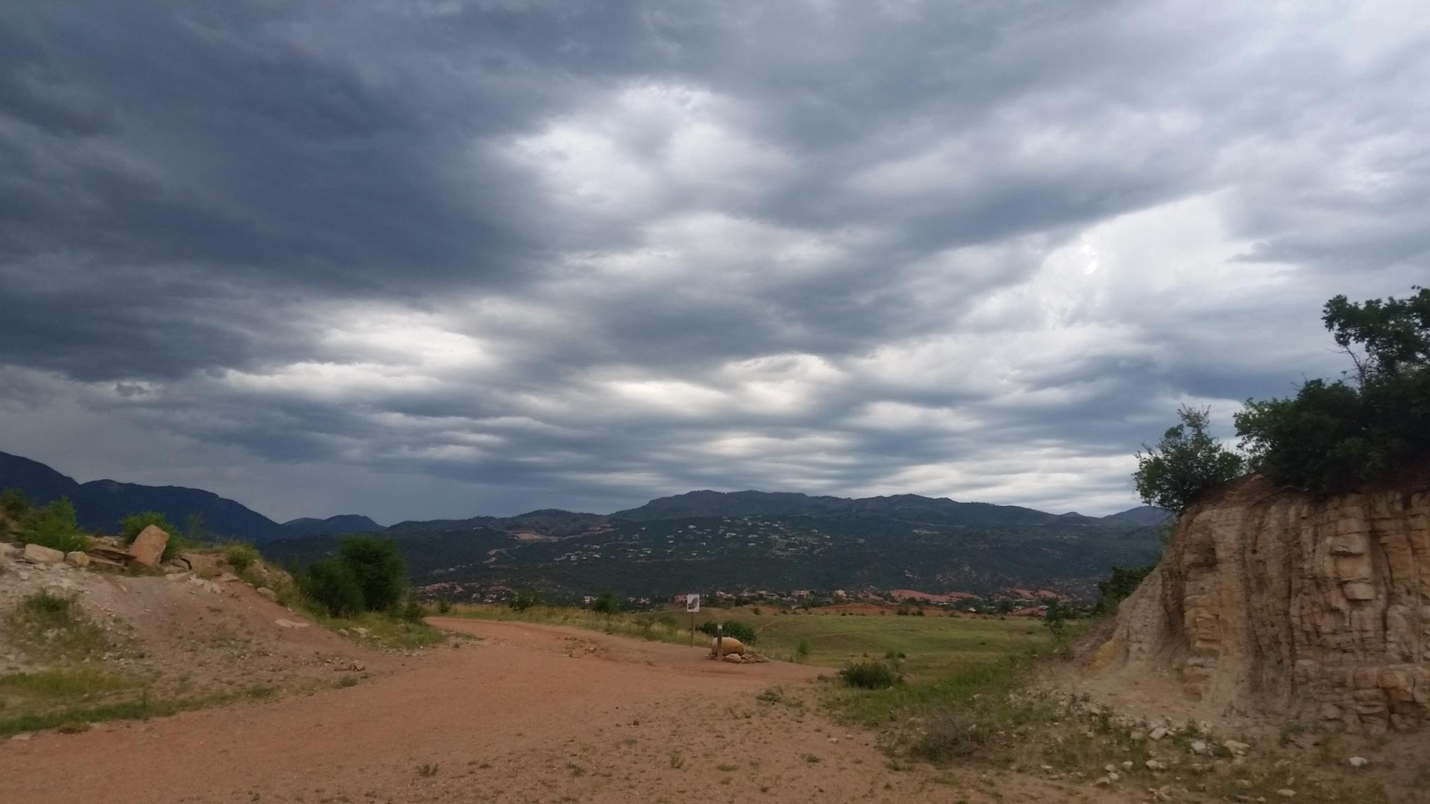 A scenic view of a dirt path winding through a landscape with rolling hills and rocky outcrops, under a dramatic sky filled with gray clouds. The background features mountains and a hint of greenery along the path, suggesting a rural and tranquil setting. Red Rock Canyon mountain bike trail.