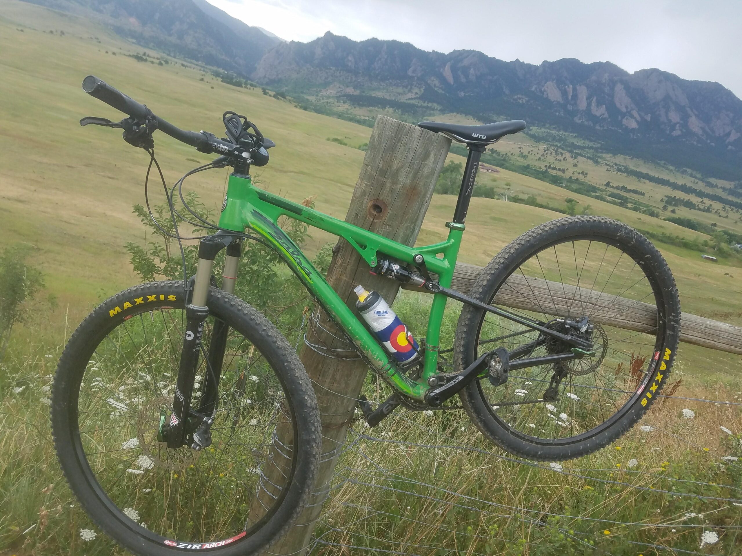 Salsa Spearfish: A green mountain bike leaning against a wooden post with a backdrop of grassy fields and mountains under a cloudy sky. The bike features black tires with yellow lettering and has a water bottle attached to the frame. Wildflowers are visible in the foreground.