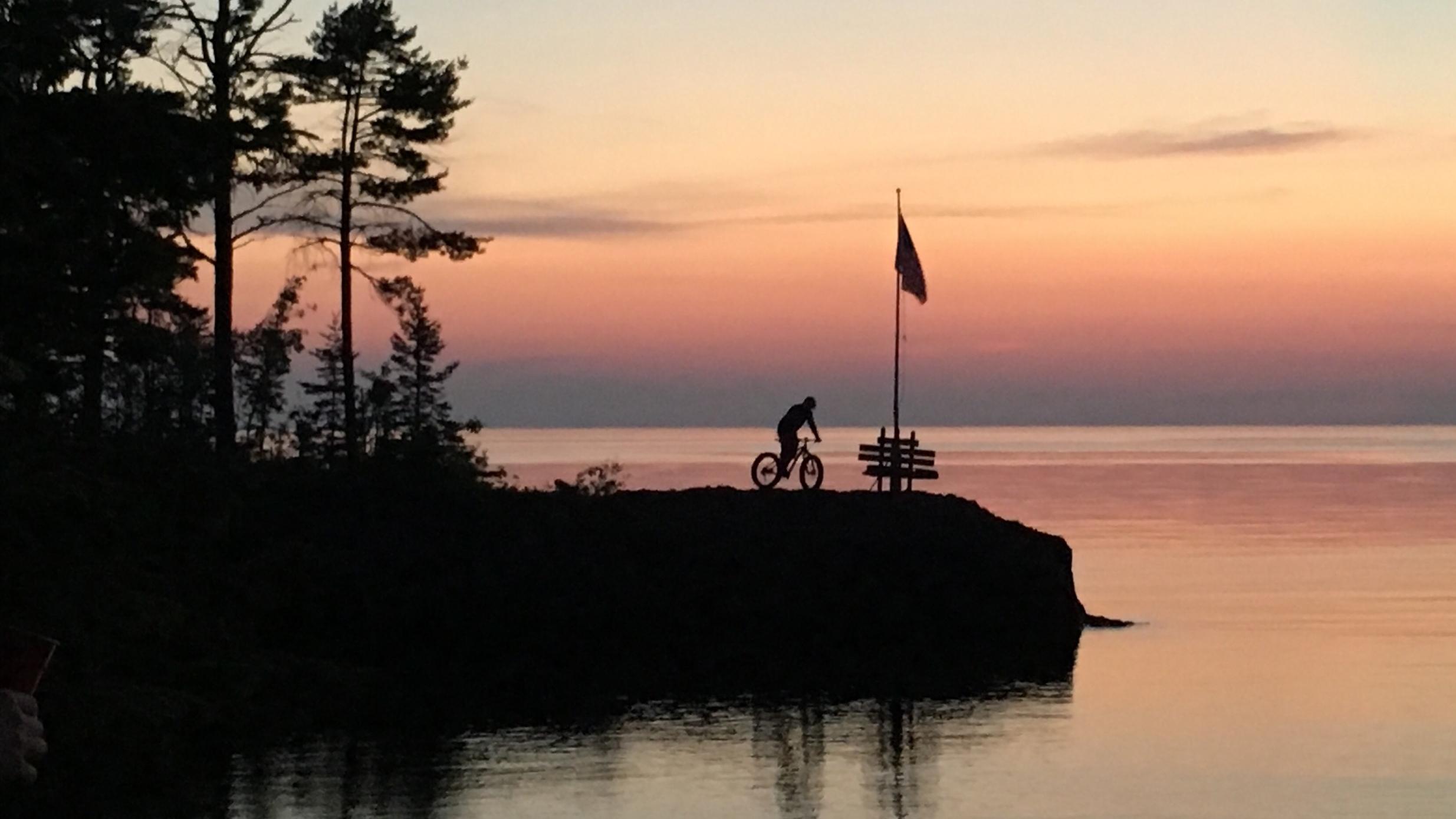 A silhouetted cyclist riding near a calm body of water during sunset, with a flag and a bench on a rocky outcrop surrounded by trees. The sky is painted in shades of pink and orange, reflecting on the water's surface. Copper Harbor Trails mountain bike trail.