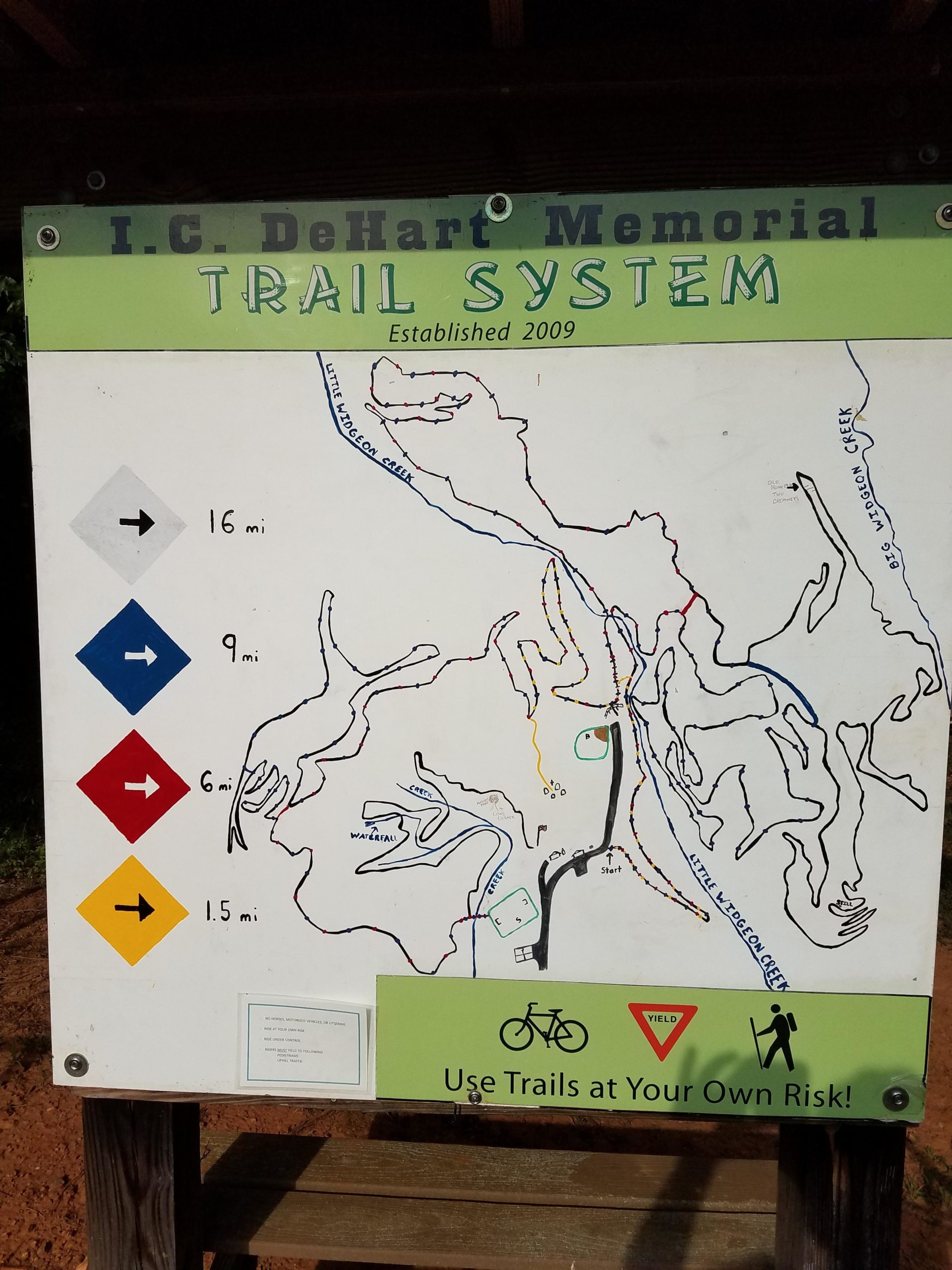 Map of the I.C. DeHart Memorial Trail System, established in 2009. The map outlines various trails with distances marked: 16 miles (blue), 9 miles (red), 6 miles (yellow), and 1.5 miles (black). It also features symbols indicating biking and hiking, along with a warning to use trails at your own risk. Woolwine Trails [Shiners Revenge] mountain bike trail.