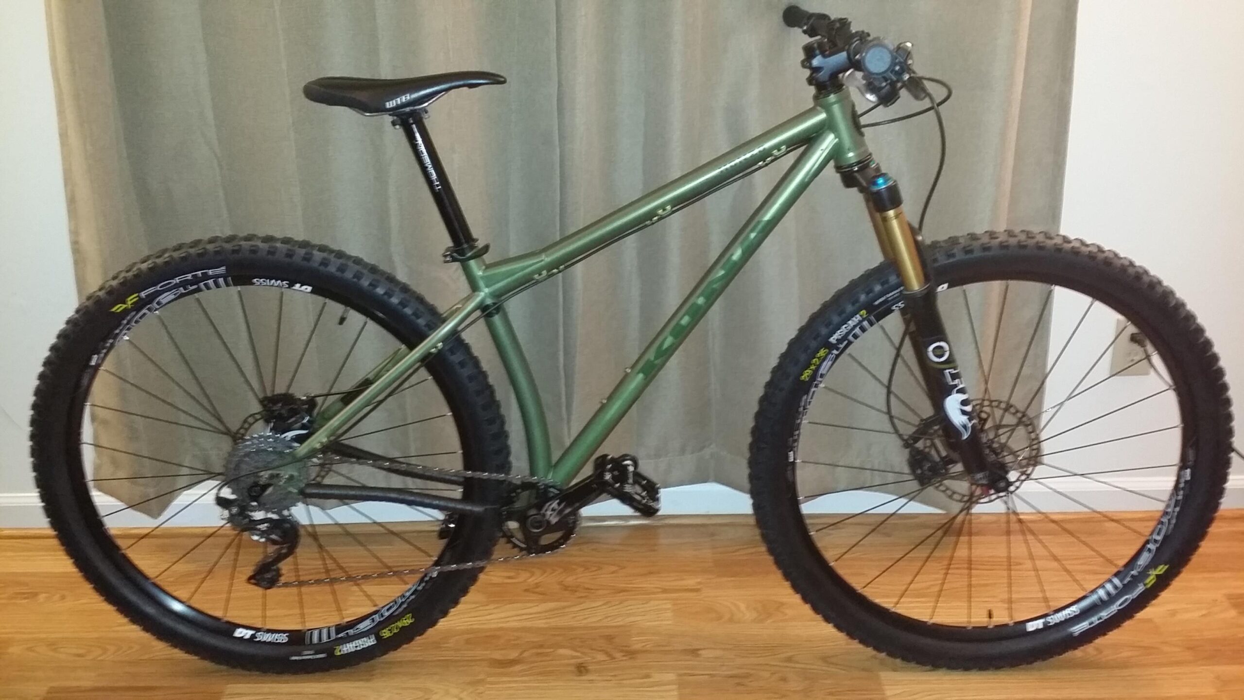 Kona Honzo: A green mountain bike with large, knobby tires is positioned upright on a wooden floor. It features a full suspension system and is equipped with disc brakes. The bike has a sleek frame design, a black seat, and handlebars with a gear shift lever. In the background, soft beige curtains create a neutral backdrop, enhancing the focus on the bike.