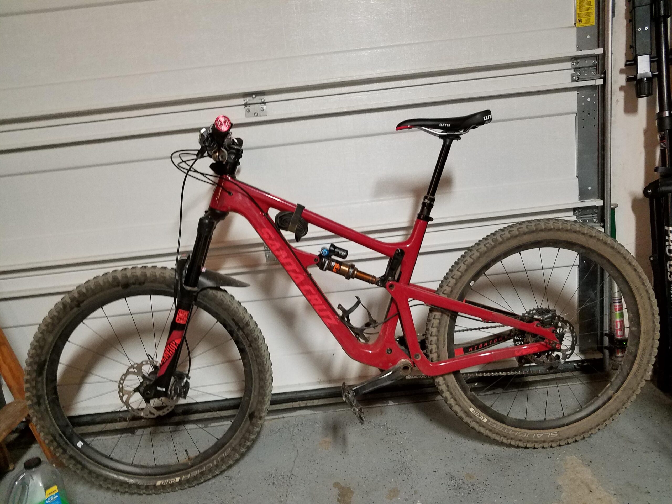 Santa Cruz Hightower 29er/27.5: A red mountain bike with a full suspension system is positioned in a garage. The bike features a prominent front fork, knobby tires, and a dropper seat post, with some visible dirt on the tires, indicating recent use. The garage door is partially closed behind it, and various tools and equipment can be seen in the background.