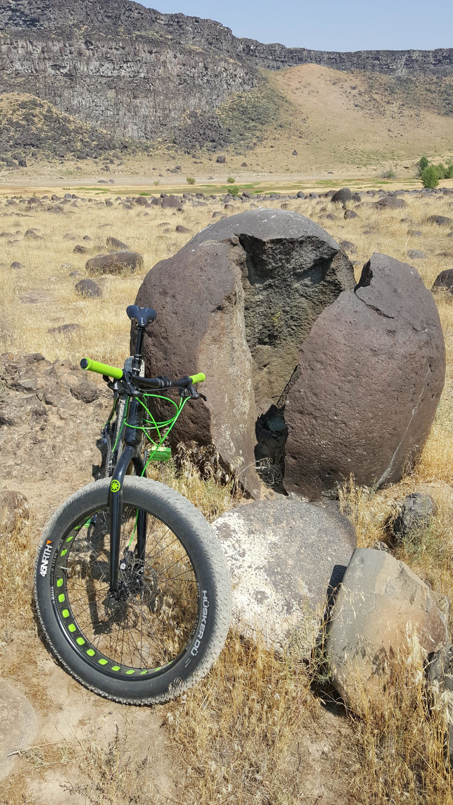 A black bicycle with green accents stands next to a large, cracked boulder in a dry, grassy landscape. The background features a rocky hill and scattered smaller rocks under a clear blue sky. Celebration river trail mountain bike trail.