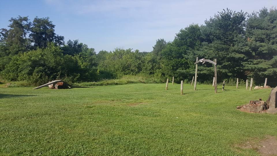 A grassy field featuring a fallen log on the left, a wooden structure resembling a swing set in the center, and several wooden posts scattered throughout, with a backdrop of trees and shrubbery under a clear blue sky. Vultures Knob mountain bike trail.