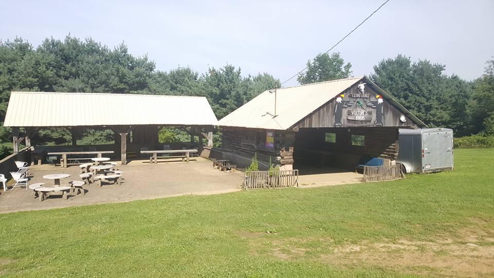 A rustic outdoor gathering space featuring two covered pavilions with wooden tables and benches, set against a backdrop of lush greenery. The structure on the right displays a sign that says "Vulture's Mine," and a trailer is parked nearby. The area appears inviting for community events or outdoor activities. Vultures Knob mountain bike trail.