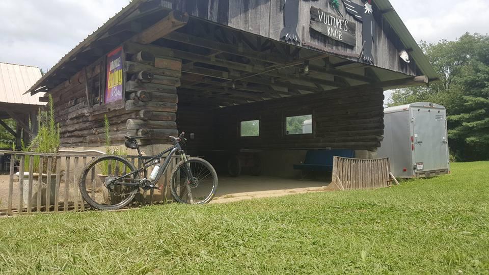 A rustic wooden structure with a sign reading "Vulture's Knob" on the front, displaying an open area with benches. A mountain bike is leaned against the building, surrounded by lush green grass and shrubs, with a gray trailer visible in the background under a partly cloudy sky. Vultures Knob mountain bike trail.
