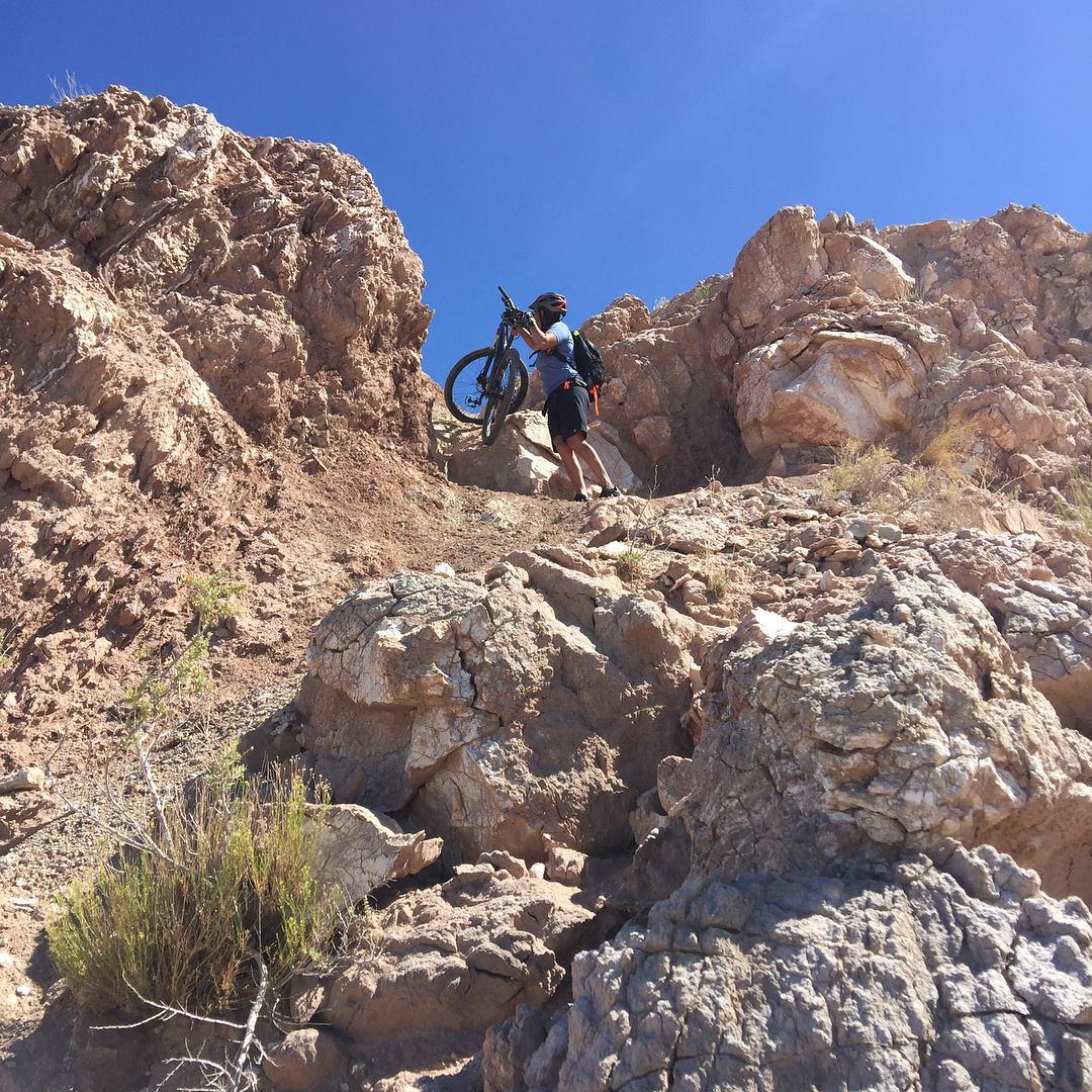 A person standing on a rocky terrain, lifting a mountain bike above their head as they navigate a steep incline. The landscape features dry, rugged rocks under a clear blue sky. White Ridge Bike Trails mountain bike trail.