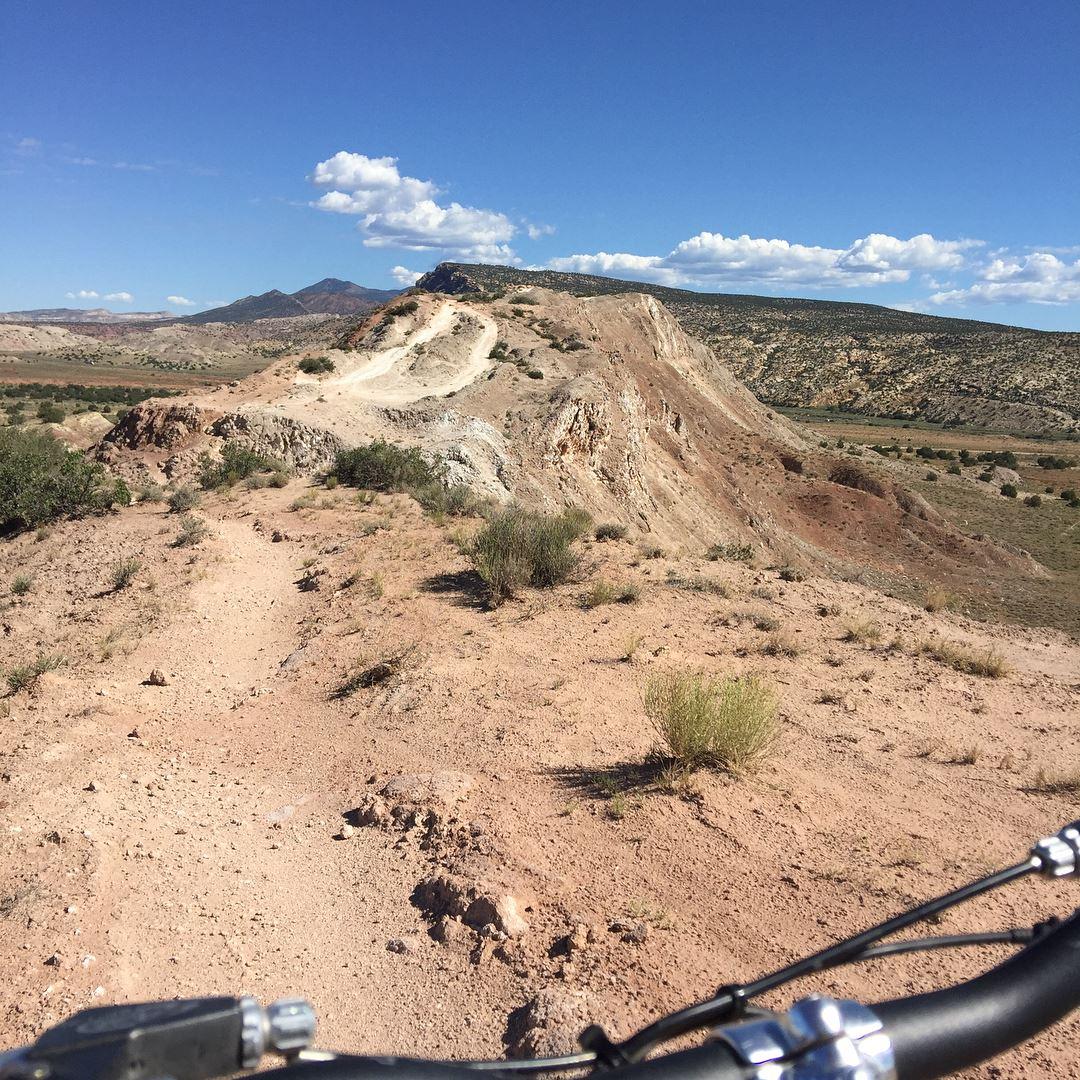 A view of a dirt trail winding through a rugged landscape with rocky hills and sparse vegetation under a clear blue sky. The image shows the perspective from a mountain bike handlebar, highlighting the path ahead that leads to a distant mountainous horizon. White Ridge Bike Trails mountain bike trail.