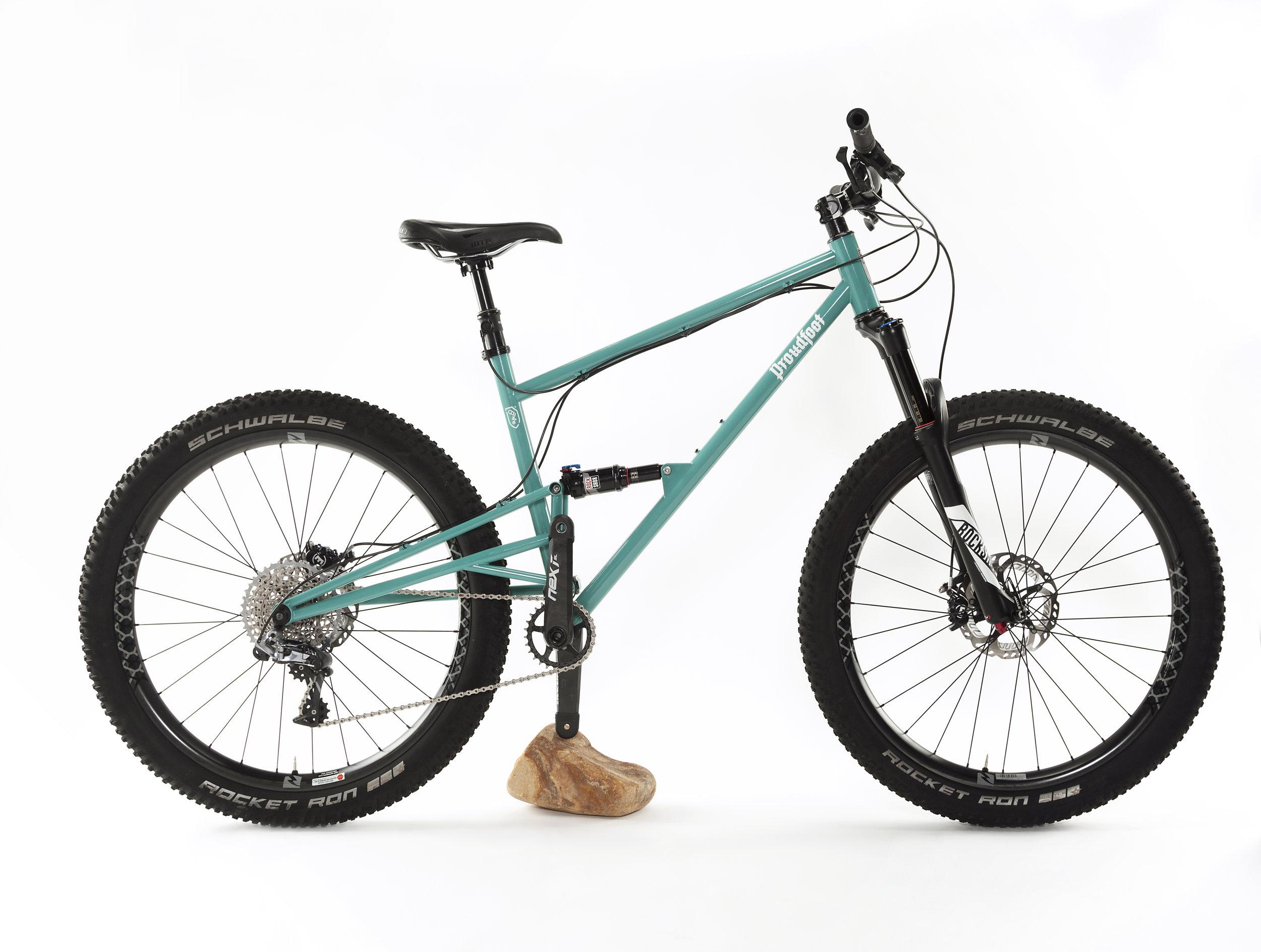 Proudfoot Cycles Primed: A teal mountain bike is positioned on a white background, featuring thick black tires with the brand name "Schwalbe" and "Rocket Ron" printed on them. The bike has a simple frame design and a prominent suspension fork at the front. A rock is placed under the bike’s rear, providing support.