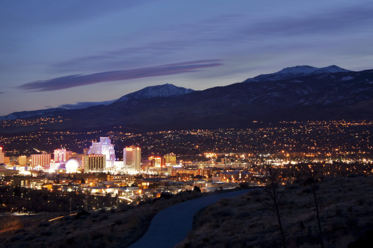 A panoramic view of a city skyline at dusk, featuring brightly lit buildings and casinos, with a backdrop of mountains and a twilight sky. The foreground includes a winding path and sparse vegetation, while the city lights reflect a vibrant nightlife atmosphere.