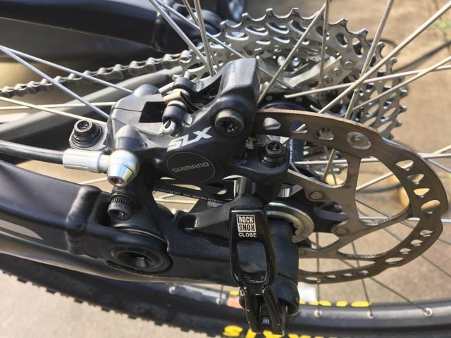 Kona Process 111: Close-up of a bicycle's rear brake system, featuring a Shimano SLX brake caliper, rotor, and surrounding components. The image highlights the detailing of the brake mechanism and the intricate arrangement of the bicycle's drivetrain and wheel.