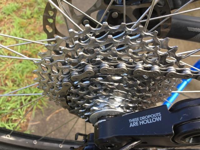 Kona Process 111: Close-up image of a bicycle gears and chain, showcasing the cassette on the rear wheel. The shiny metal components are clearly visible, along with a portion of the bicycle's dropout with the text "THESE DROPOUTS ARE HOLLOW." The background features green grass and a concrete surface.