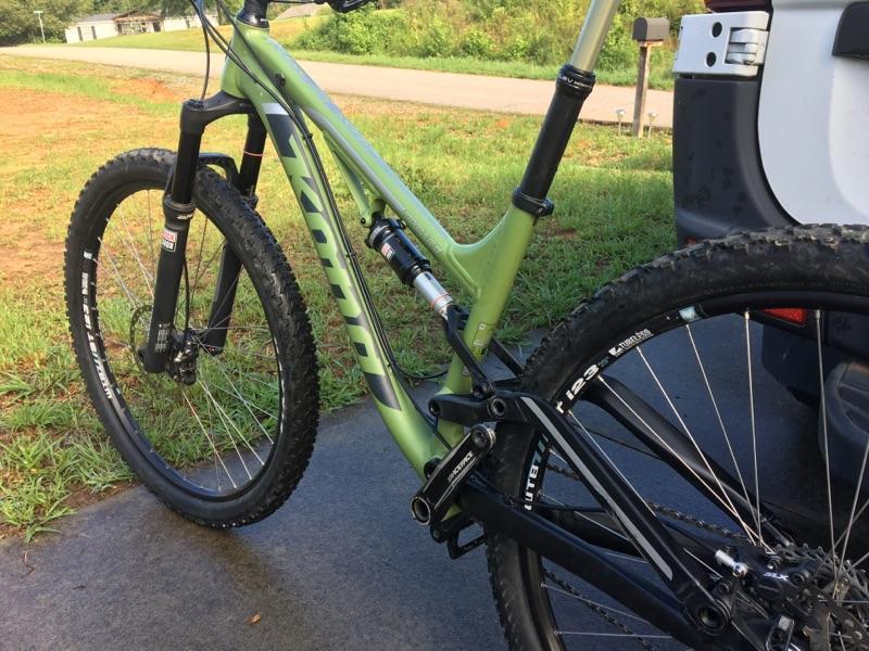 Kona Process 111: A close-up view of a green mountain bike, showcasing the front fork, wheel, and part of the frame. The bike is parked on a paved surface with grass in the background, and a vehicle is partially visible to the right. The tires have visible tread patterns, indicating they are suited for off-road use.