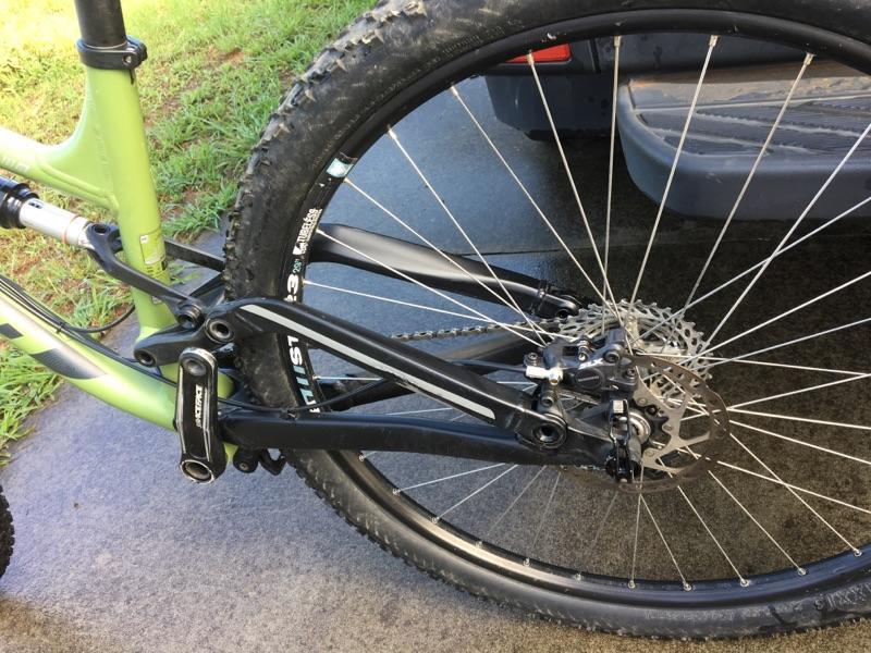 Kona Process 111: Close-up view of the rear wheel and drivetrain of a mountain bike, showcasing the black rear suspension components, disc brake, and gear system, with a grassy area and part of a vehicle in the background.
