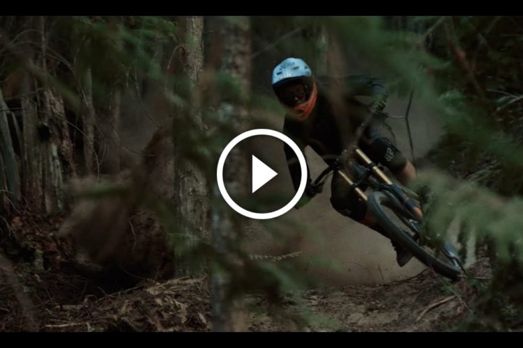A mountain biker navigating a winding dirt trail through a forest, with trees and foliage surrounding the path. Dust is kicked up as the rider leans into a turn, wearing protective gear and a helmet.