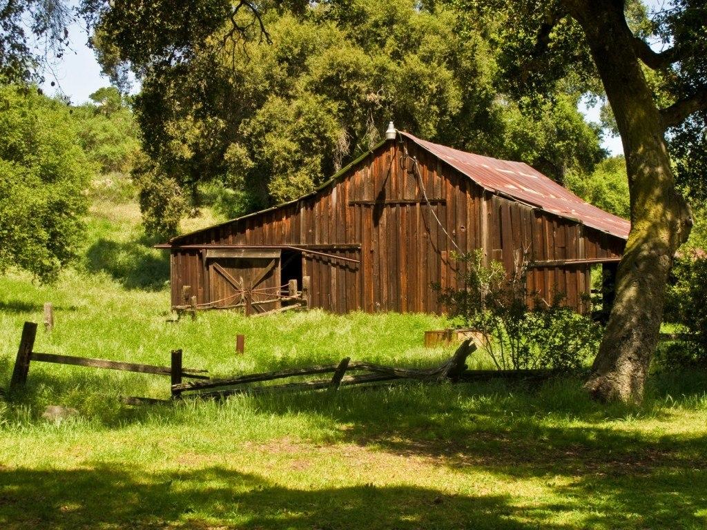 A rustic wooden barn with a weathered roof, surrounded by lush green grass and trees. A wooden fence is partially visible in the foreground. The scene evokes a serene countryside atmosphere. Daley Ranch mountain bike trail.