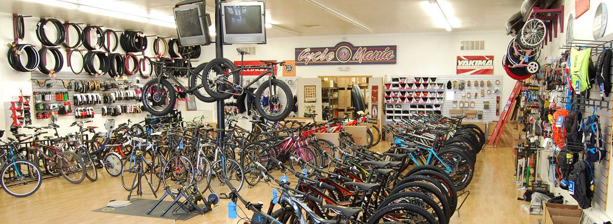 A spacious bike shop interior filled with a variety of bicycles and cycling accessories. The walls are lined with bike wheels, helmets, and gear. Multiple bikes are displayed prominently on the sales floor, while bike maintenance equipment can be seen in the center. The shop is well-lit, creating an inviting atmosphere for customers.