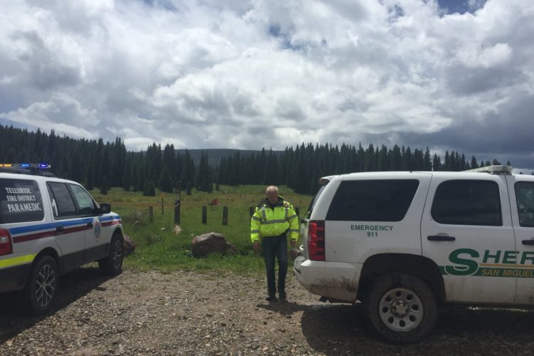 A paramedic vehicle from the Telluride Fire District and a Sheriff’s vehicle from San Miguel are parked in a grassy, wooded area. A uniformed officer is standing near the sheriff's vehicle, with trees and a cloudy sky in the background, indicating a remote location.