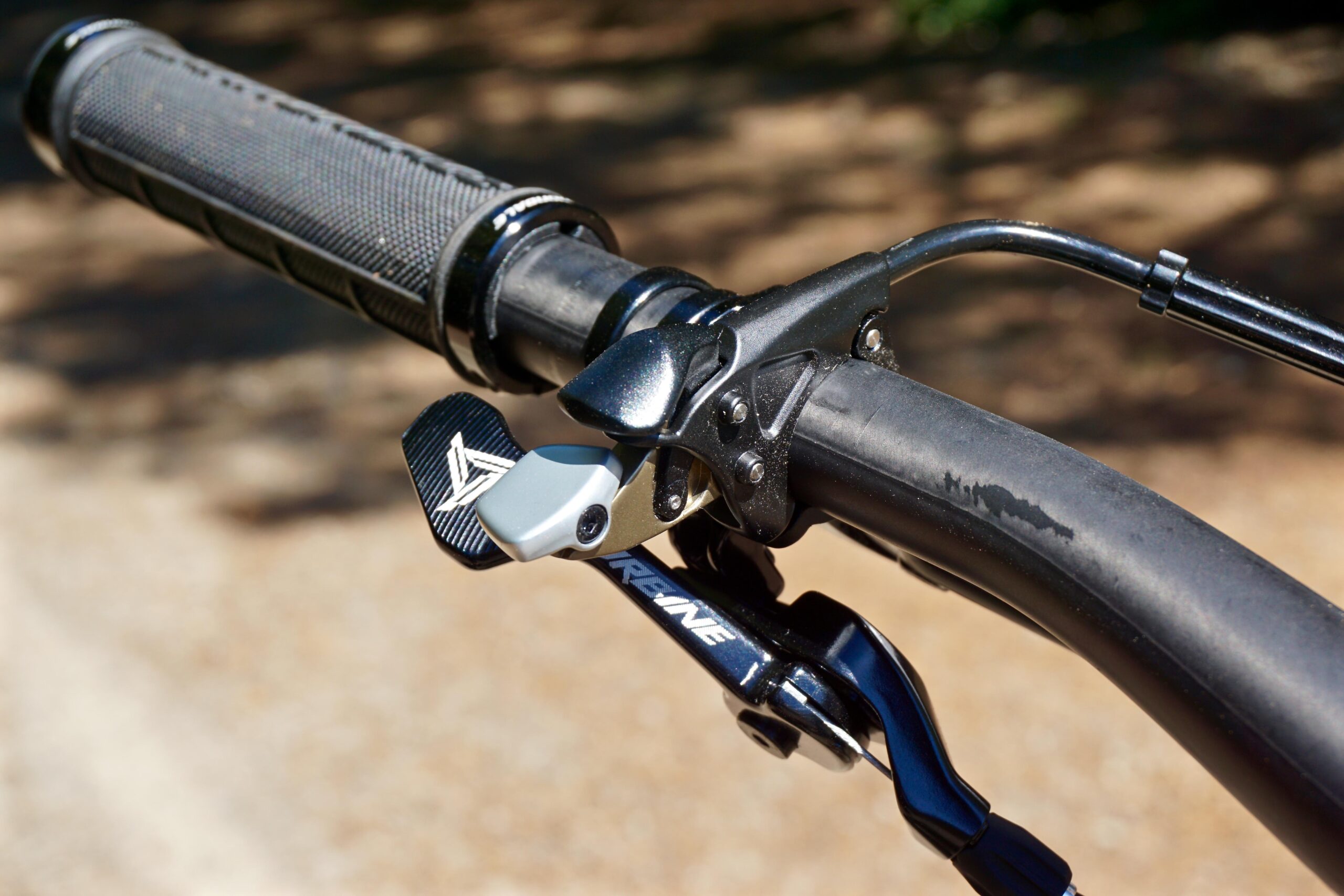 Cannondale Trigger: Close-up view of a bicycle handlebar featuring a textured black grip and a silver brake lever, with a shiny black and silver shifter attached. The background shows a blurred out, gravel path.