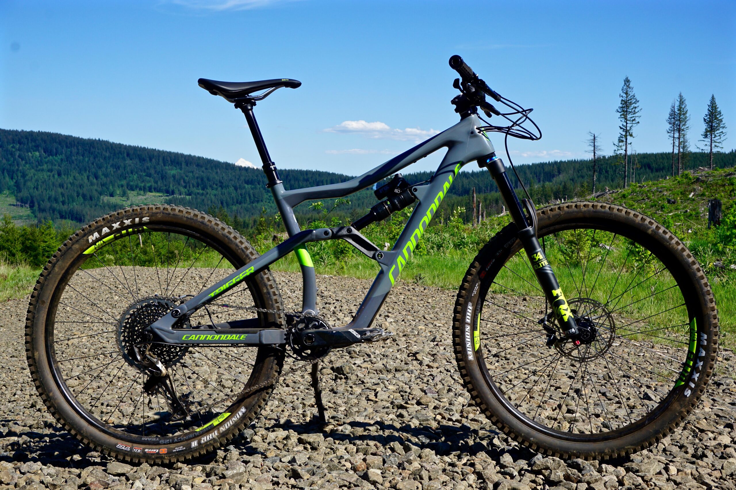 Cannondale Trigger: A mountain bike with a grey and green frame is positioned on a gravel path, surrounded by lush greenery and distant hills. The bike features modern components, including thick tires with Maxxis branding, and a front suspension fork, ideal for off-road cycling. The clear blue sky in the background enhances the outdoor setting.