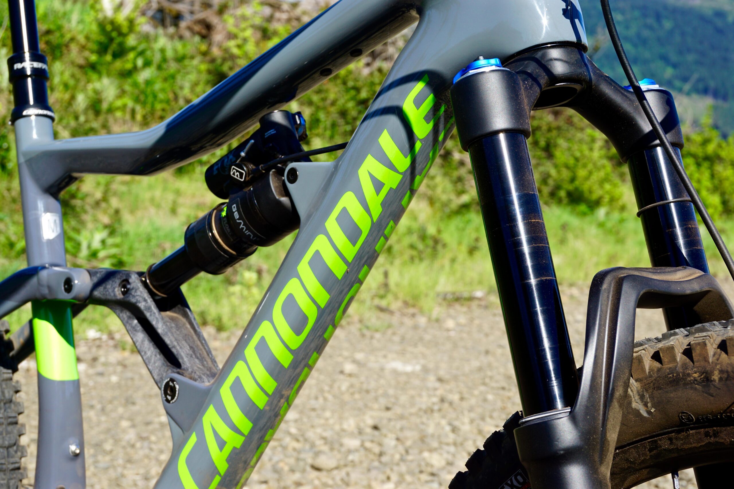 Cannondale Trigger: Close-up of a gray Cannondale mountain bike frame, highlighting the suspension system with a black shock absorber and a front fork. The bike features bright green lettering on the frame and is positioned against a blurred natural background with greenery, indicating an outdoor setting.