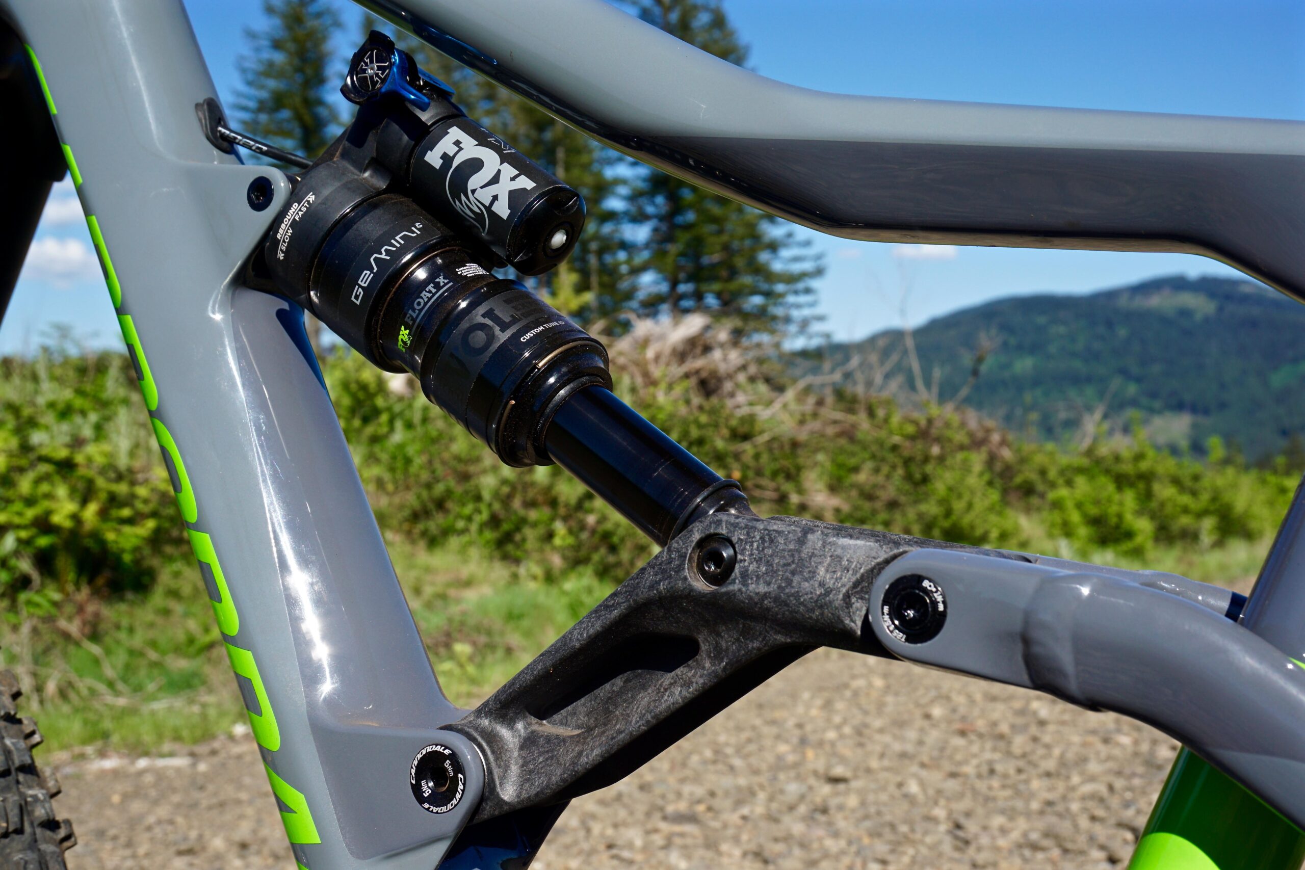 Cannondale Trigger: Close-up view of a mountain bike's rear shock absorber, showcasing the FOX suspension system mounted on a sleek gray frame with bright green accents. The background features a lush landscape with trees and hills under a clear blue sky.