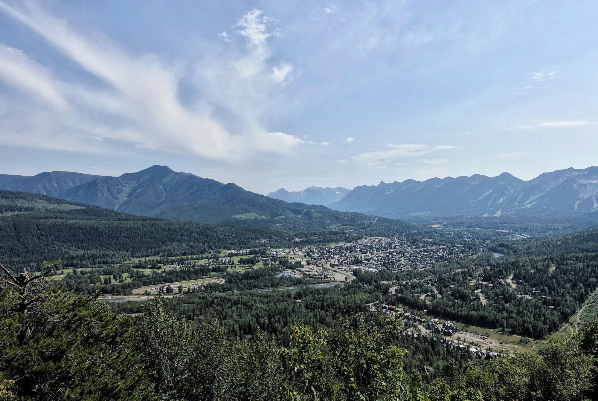 A panoramic view of a valley surrounded by mountains, featuring lush greenery and a small town nestled in the foreground. The sky is partly cloudy, showcasing a serene and picturesque landscape. Swine Flu Loop mountain bike trail.