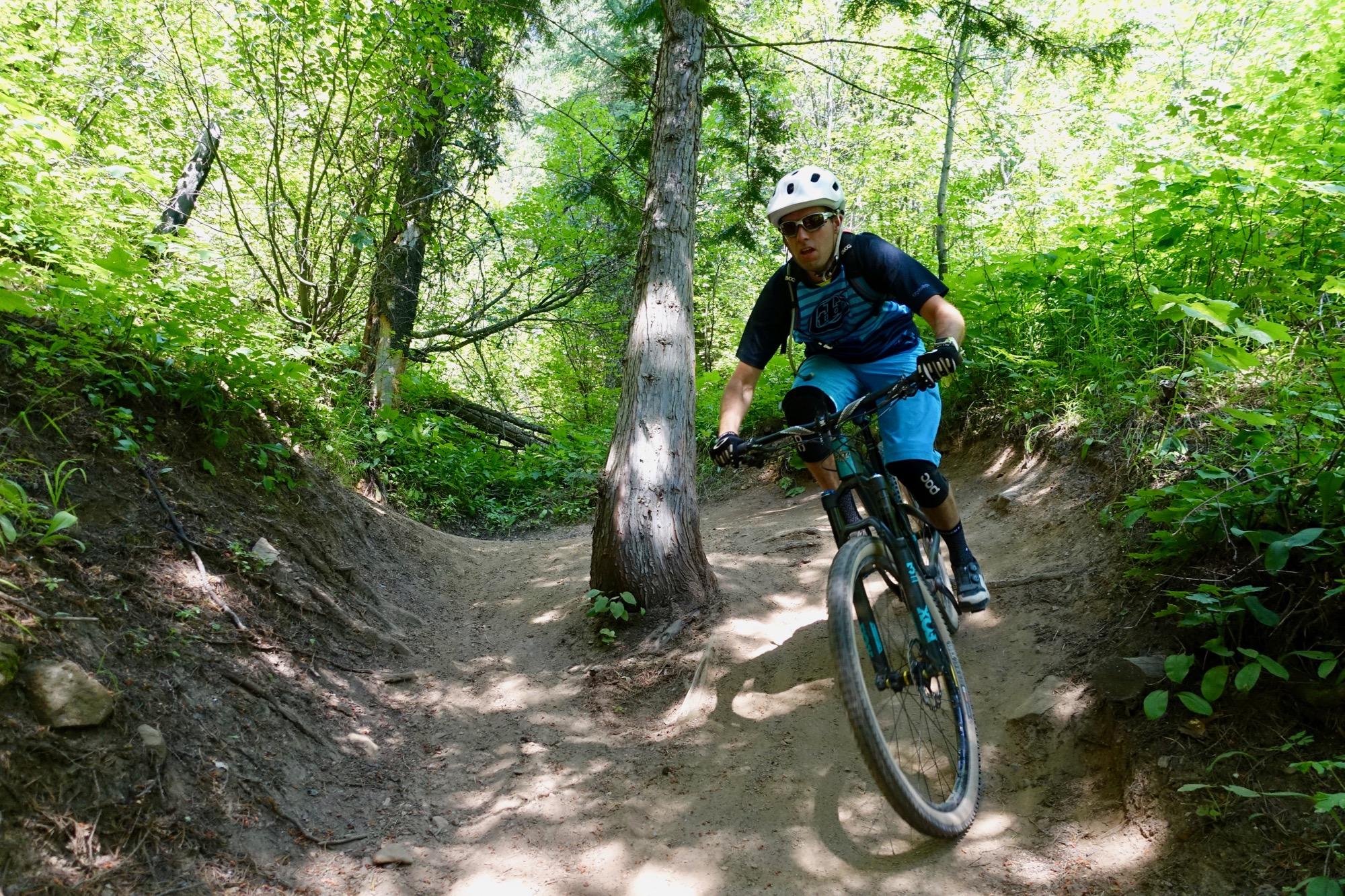 A mountain biker navigating a dirt trail surrounded by lush green vegetation and trees, wearing a white helmet, sunglasses, and a blue athletic outfit. The path curves around a tree, emphasizing the dynamic nature of the ride. Swine Flu mountain bike trail.