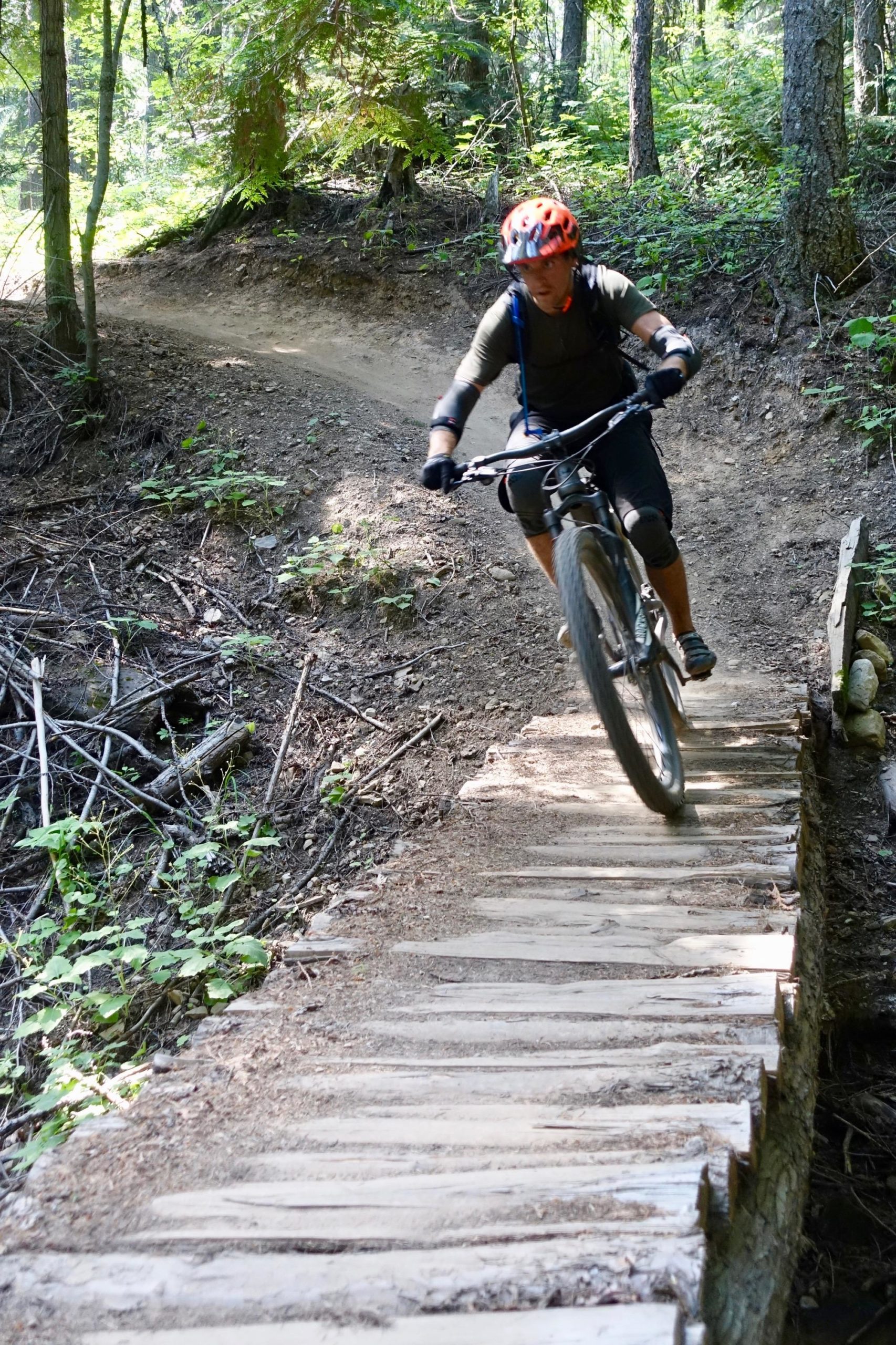 A mountain biker wearing a helmet and protective gear navigates a wooden bridge on a woodland trail, surrounded by lush greenery. The cyclist is in an action pose as they ride over the bridge, which is set against a backdrop of trees and dirt paths. Swine Flu Loop mountain bike trail.