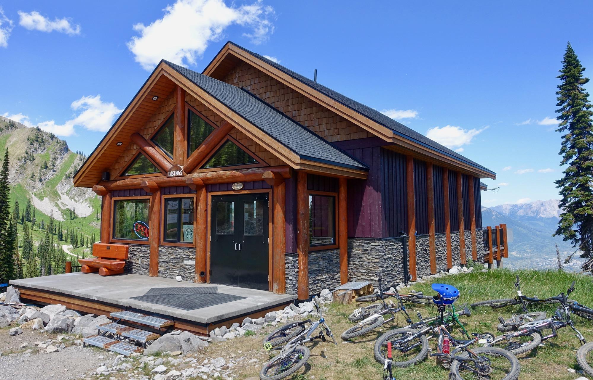 A rustic lodge with wooden beams and stone accents, featuring large windows and an "Open" sign. In front, several mountain bikes are parked on the grass, with a beautiful mountainous landscape and blue sky in the background. Rumplestumpskin mountain bike trail.
