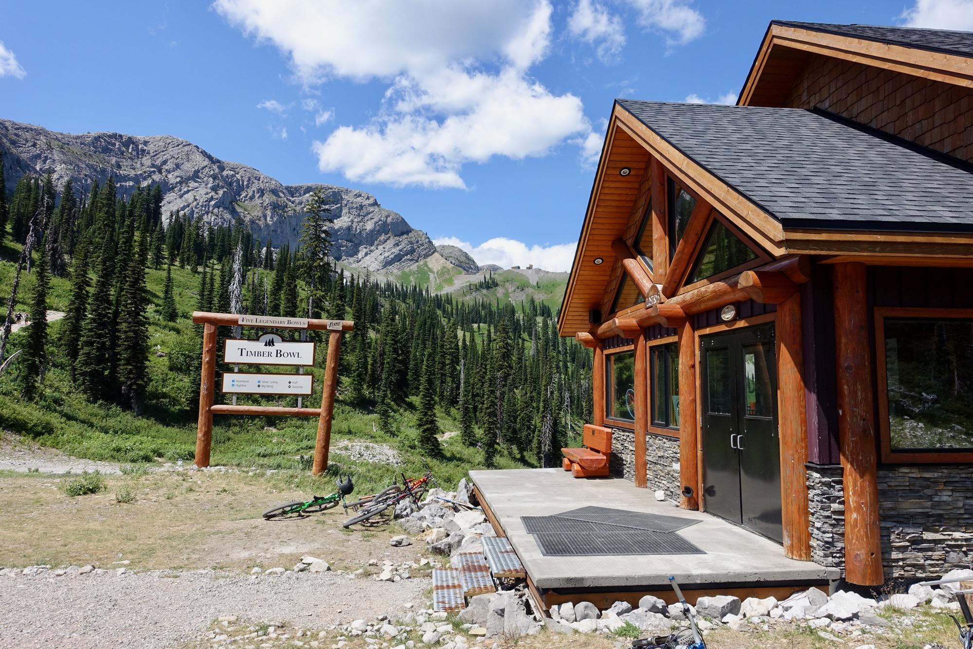A rustic building with a wooden exterior and large windows, located in a mountain setting. In front, a wooden sign reads "Timber Bowl," indicating the area. Bicycles are parked nearby, with green pine trees and rocky mountains in the background under a bright blue sky dotted with clouds. Rumplestumpskin mountain bike trail.