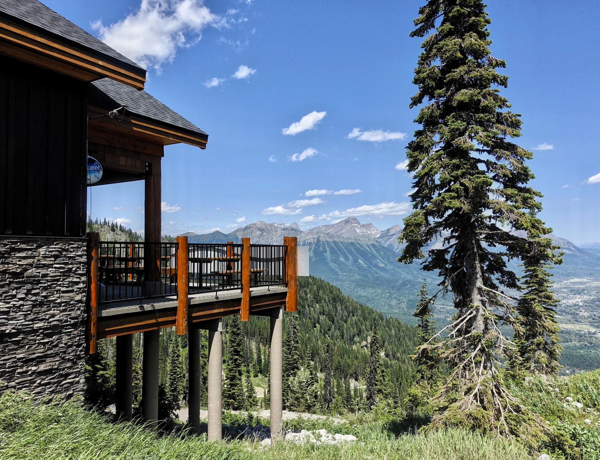 A mountain lodge with a wooden deck overlooks a scenic valley, surrounded by tall evergreen trees and rugged mountain peaks under a clear blue sky with fluffy white clouds. The lodge features dark wood and stone construction, with a prominent railing on the deck. Rumplestumpskin mountain bike trail.