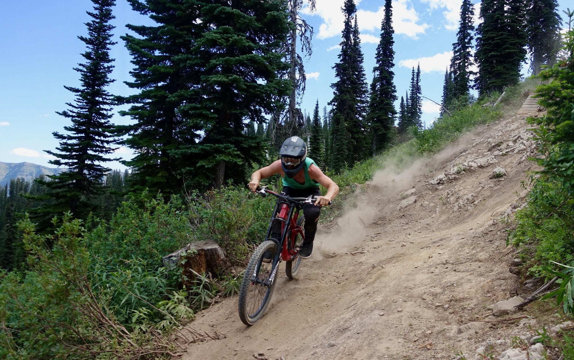 A person wearing a helmet and protective gear rides a mountain bike down a dusty trail surrounded by tall evergreen trees. The rider is focused and leaning forward, with a trail of dust being kicked up behind them as they navigate the downhill path. In the background, there are mountains visible under a bright blue sky with scattered clouds. Rumplestumpskin mountain bike trail.