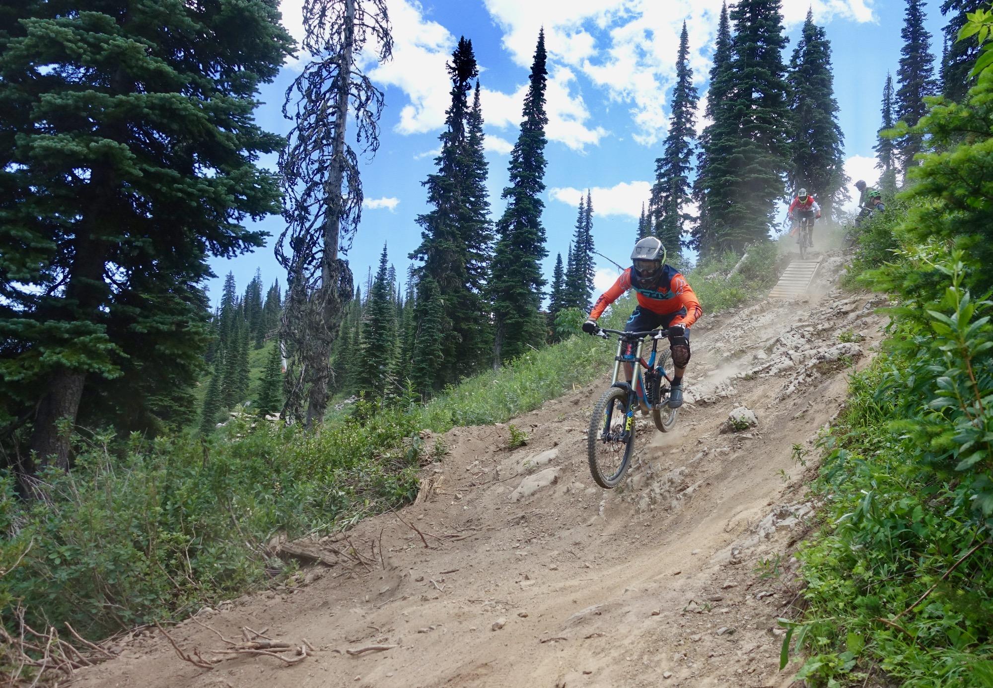 A mountain biker in an orange jersey navigates a dusty downhill trail surrounded by tall pine trees under a blue sky with scattered clouds. Dust is kicked up as the rider leans into the turn, showcasing the excitement of mountain biking in a natural setting. Rumplestumpskin mountain bike trail.
