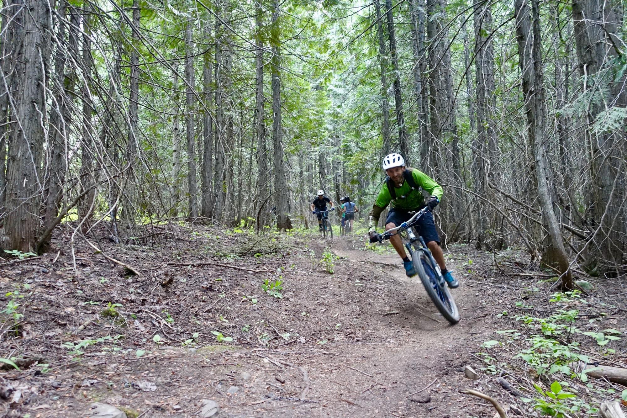 Mountain bikers navigating a winding dirt trail through a dense forest, surrounded by tall trees and underbrush. One rider, dressed in a green jacket and white helmet, leans into a turn, while another cyclist follows behind in the background. Mount Fernie Provincial Park mountain bike trail.