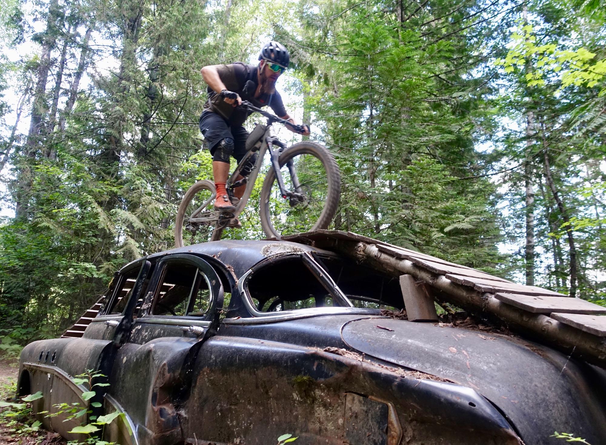 A mountain biker performs a trick while riding on top of an abandoned car in a forested area. The bike and rider are in mid-air, showcasing the bike's suspension. Surrounding the scene are tall trees and lush greenery, emphasizing a natural outdoor setting. Red Sonja mountain bike trail.