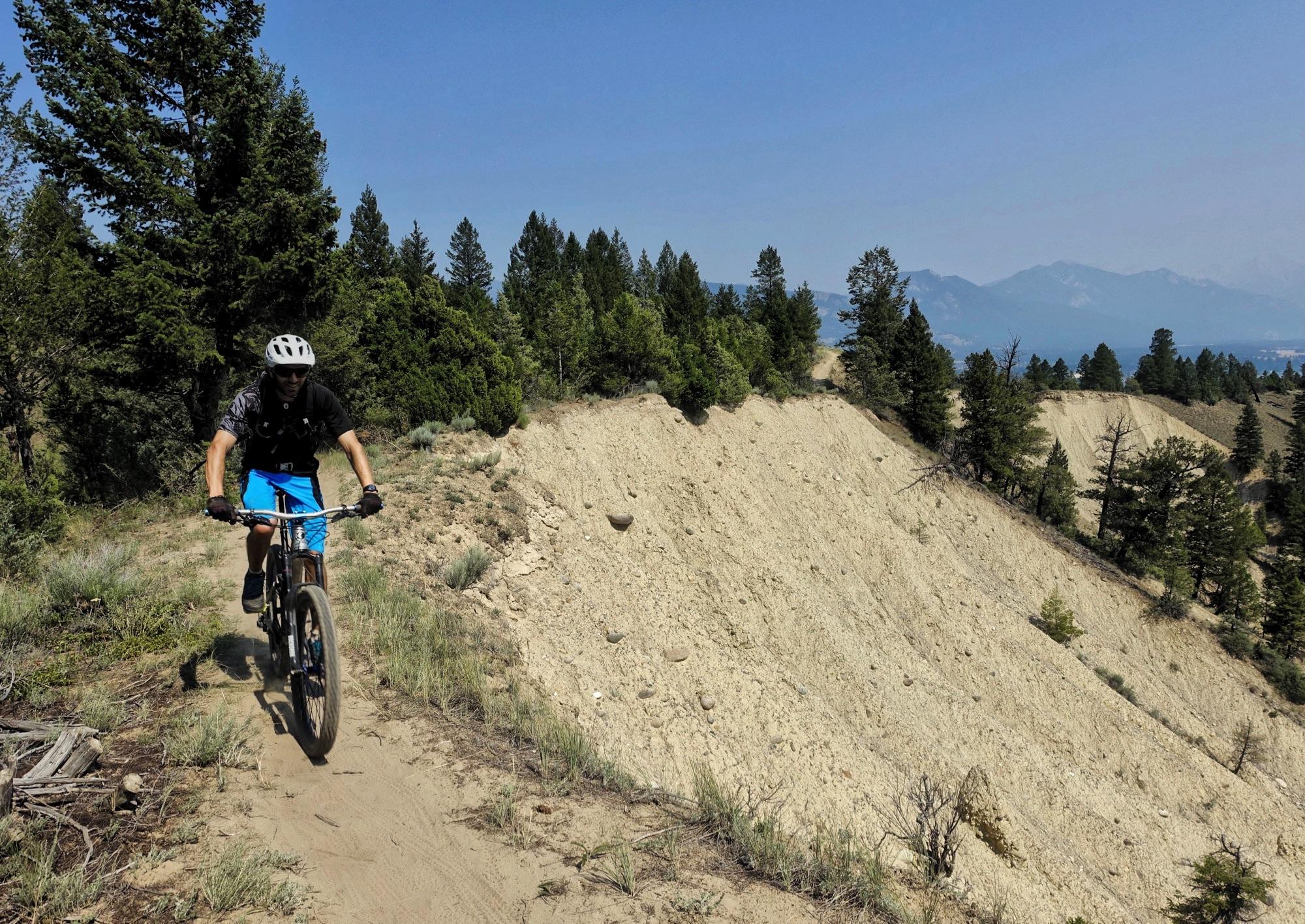 A mountain biker rides along a narrow dirt trail surrounded by trees and a steep slope on one side. The landscape features a mix of greenery and rocky terrain under a clear blue sky, with distant mountains visible in the background. Lake Lillian mountain bike trail.
