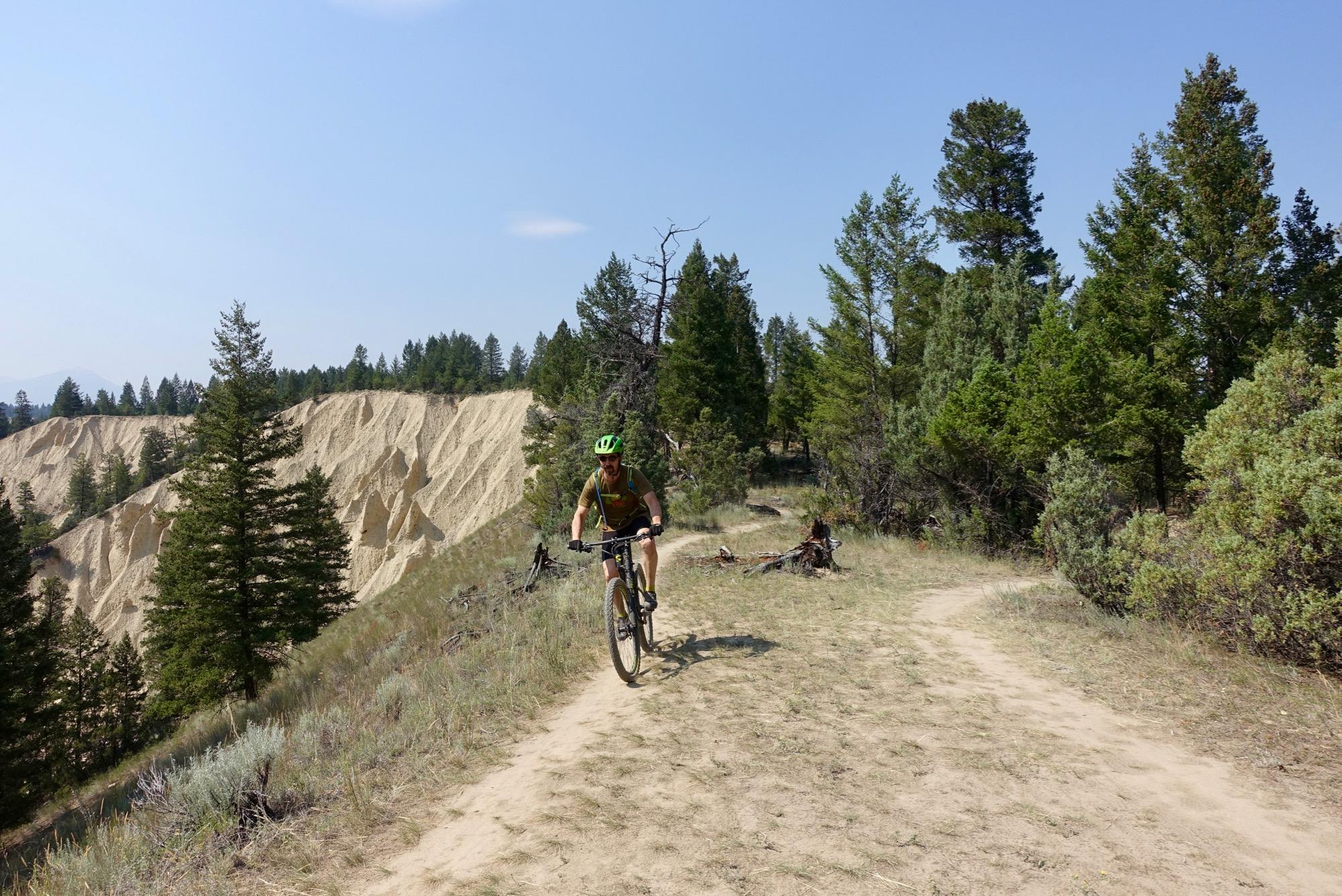 A mountain biker riding along a sandy trail in a forested area, with pine trees in the background and a sandy hillside to the left. The sky is clear and blue, indicating a sunny day. Lake Lillian mountain bike trail.