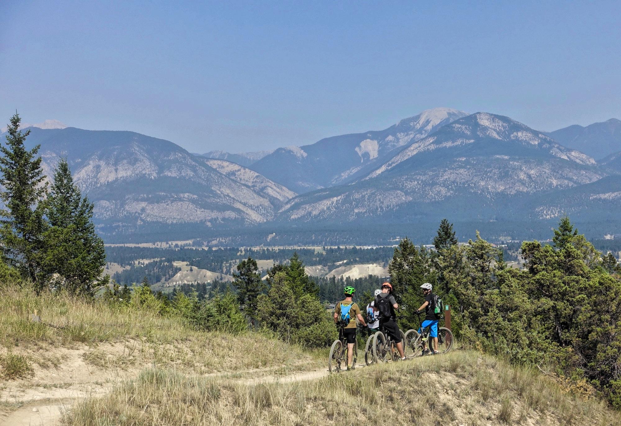 A group of four mountain bikers stands on a dirt trail, overlooking a scenic view of mountains and valleys under a clear blue sky. Lush greenery surrounds the trail, adding to the outdoor adventure atmosphere. Lake Lillian mountain bike trail.