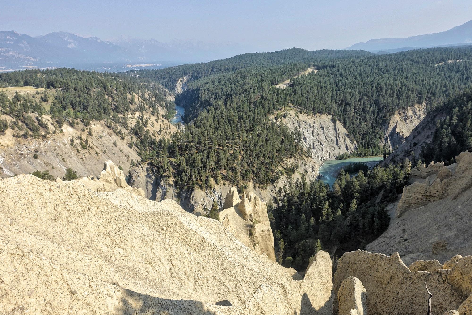 A panoramic view of a steep, rugged landscape featuring a mix of rocky cliffs and dense pine forests, with a winding river visible in the valley below. The scene is set against a backdrop of distant mountains under a clear sky. The foreground displays textured soil formations, while the lush green trees create a natural contrast with the earthy tones of the cliffs. Lake Lillian mountain bike trail.