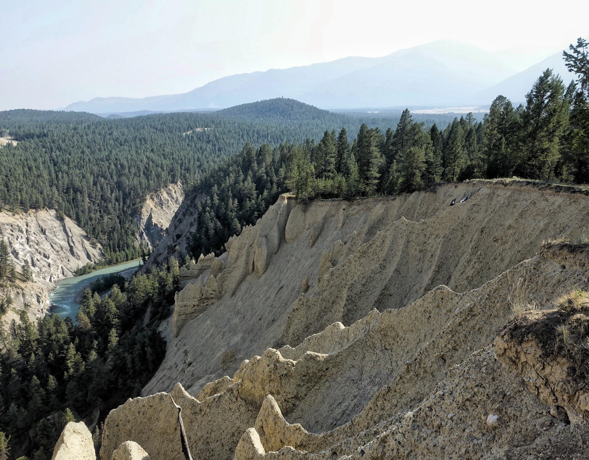 A panoramic view of a mountainous landscape featuring steep, rugged cliffs with distinct layered formations. Below, a winding river flows through a dense forest of evergreen trees. The scene is framed by distant mountains under a clear sky. Lake Lillian mountain bike trail.