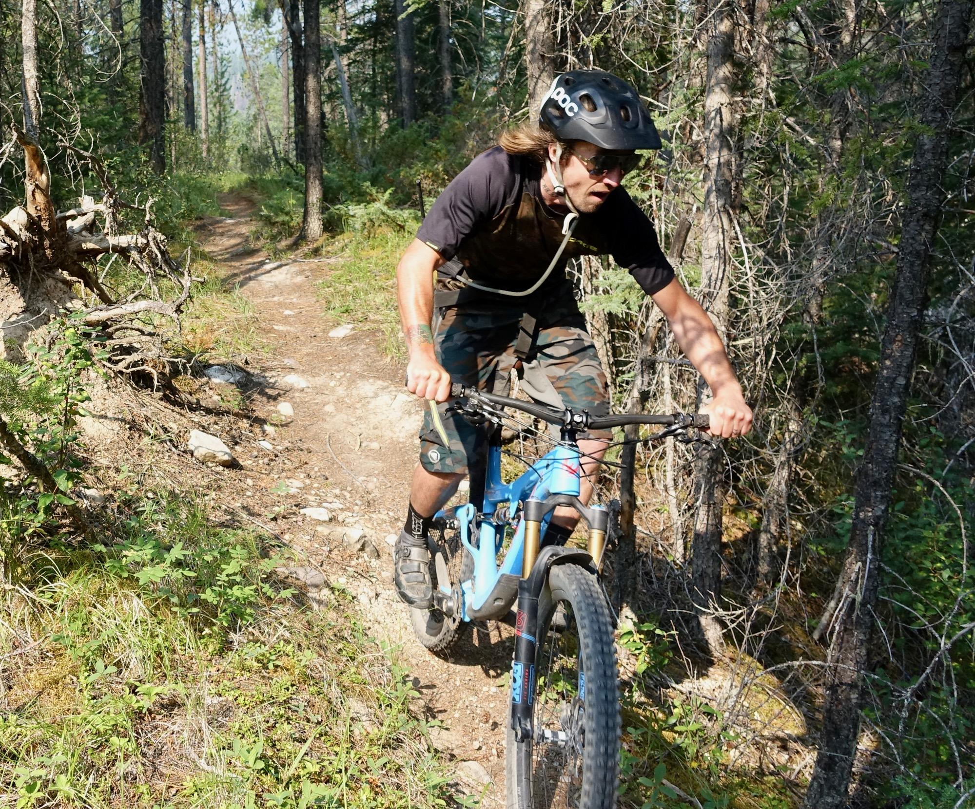 A mountain biker navigating a narrow dirt trail in a forested area, wearing a helmet and sunglasses. The bike is blue, and the rider is focused on the path ahead, surrounded by trees and greenery. Nipika Mountain Resort mountain bike trail.