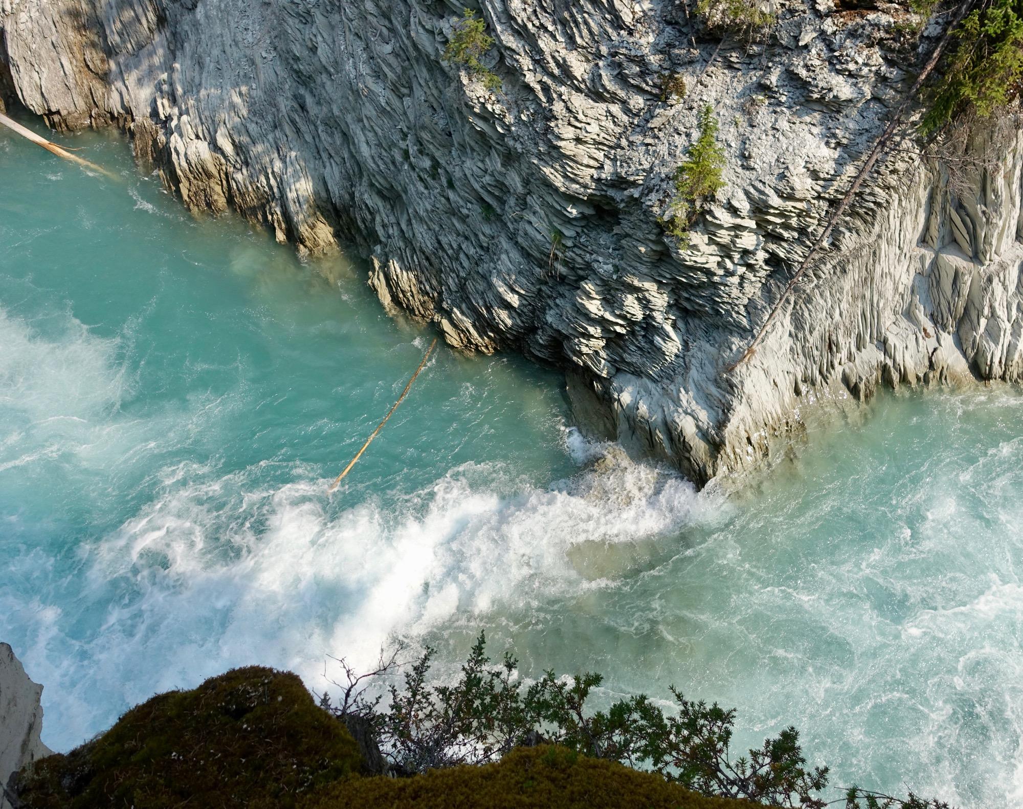 Aerial view of a turquoise river flowing over rocky terrain, with waves crashing against the cliffs. The scene includes patches of green vegetation along the water's edge and fallen logs partially submerged in the river. Nipika Mountain Resort mountain bike trail.