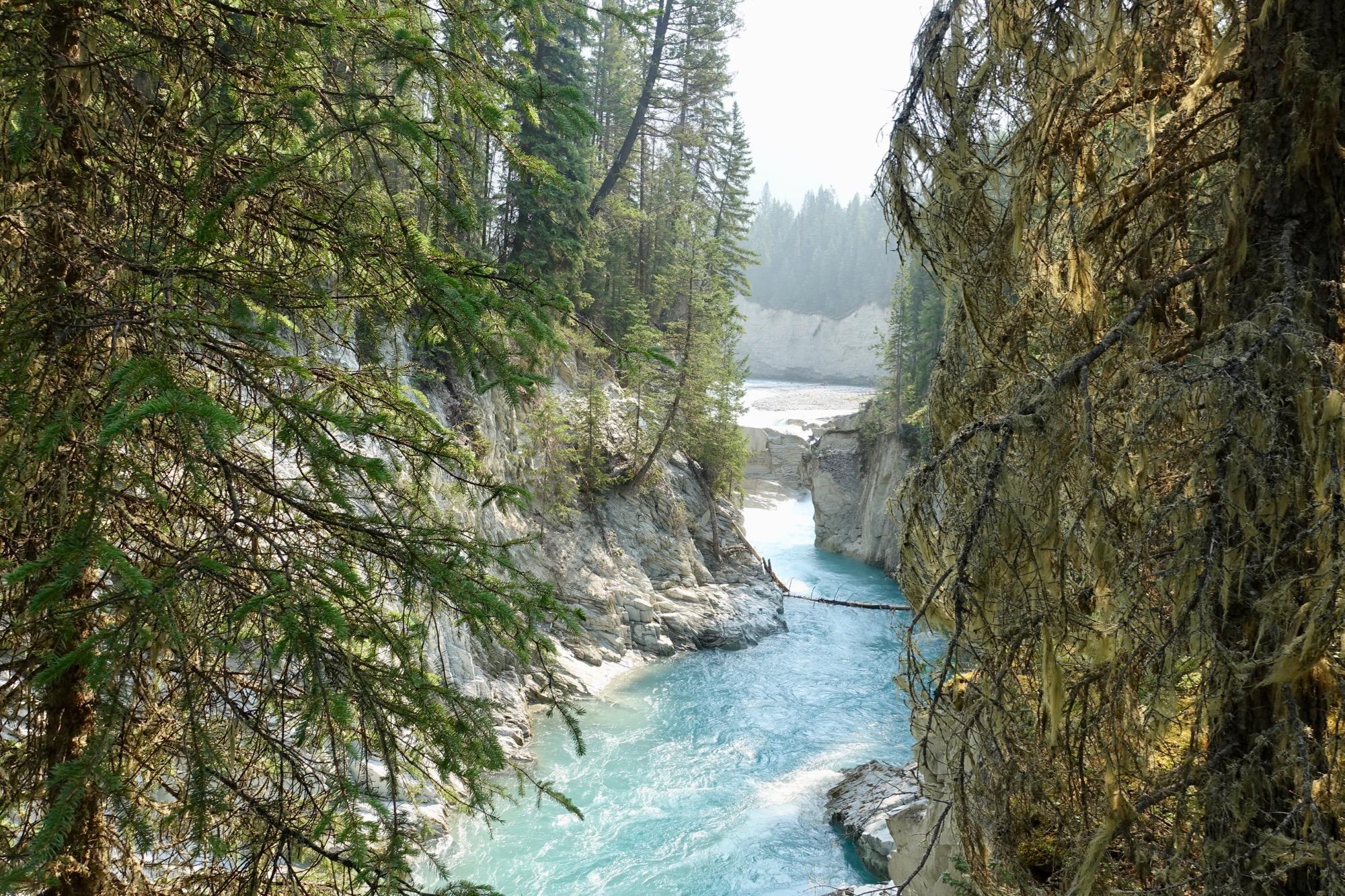 A serene view of a river winding through rocky cliffs, surrounded by lush green trees. The water appears turquoise, contrasting beautifully with the gray rocks and the vibrant foliage. The landscape is gently illuminated by soft light, suggesting a tranquil and untouched natural environment. Nipika Mountain Resort mountain bike trail.