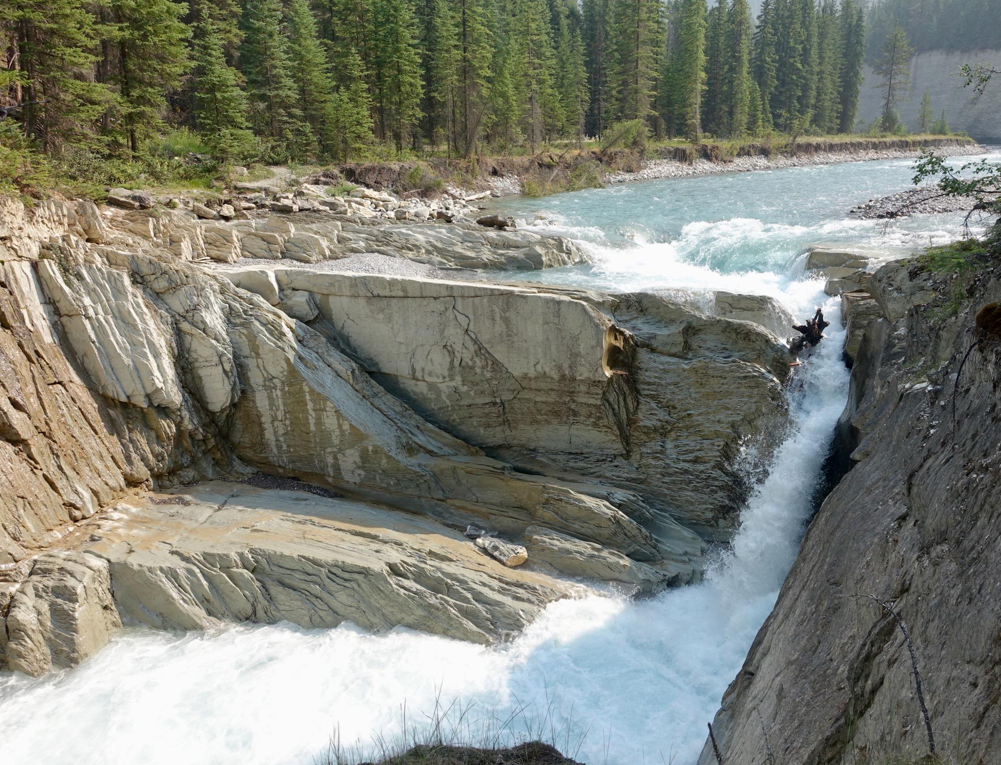 A rocky riverbank with smooth, layered stone formations cascading into a rushing turquoise river. Dense coniferous trees line the background, creating a natural, serene landscape. The water flows rapidly over the rocks, generating white foamy waves. Nipika Mountain Resort mountain bike trail.