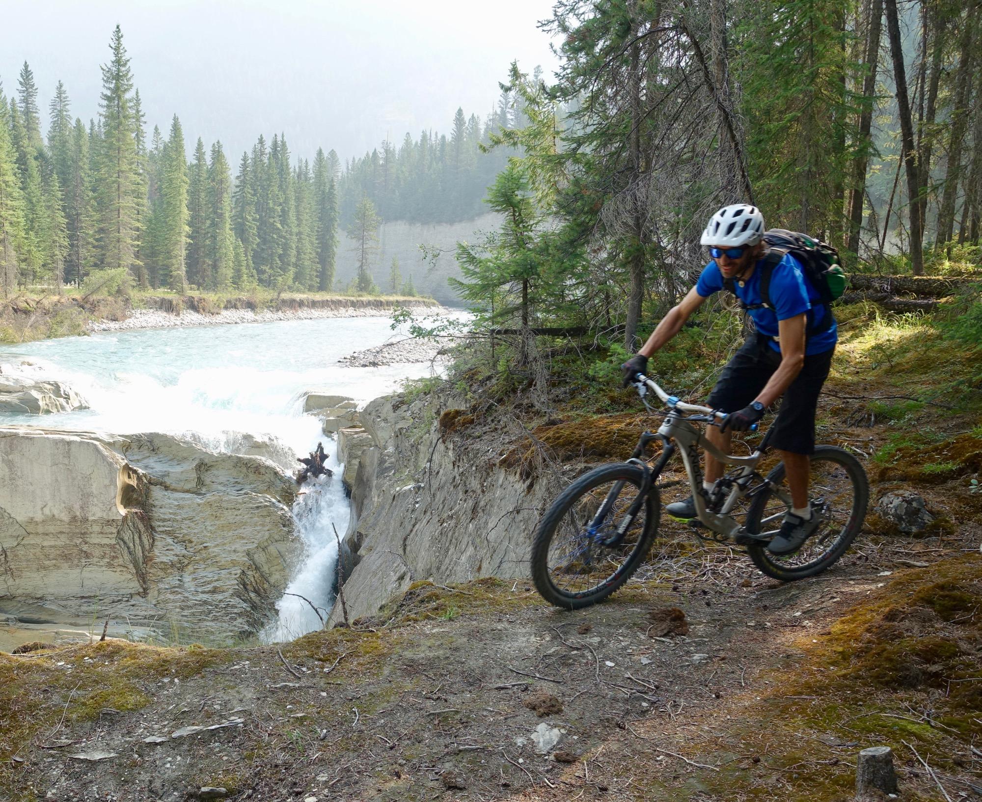 A person riding a mountain bike along a narrow trail near a river, surrounded by tall pine trees and rocky terrain. Water cascades over rocks in the background, creating a scenic natural landscape. Nipika Mountain Resort mountain bike trail.