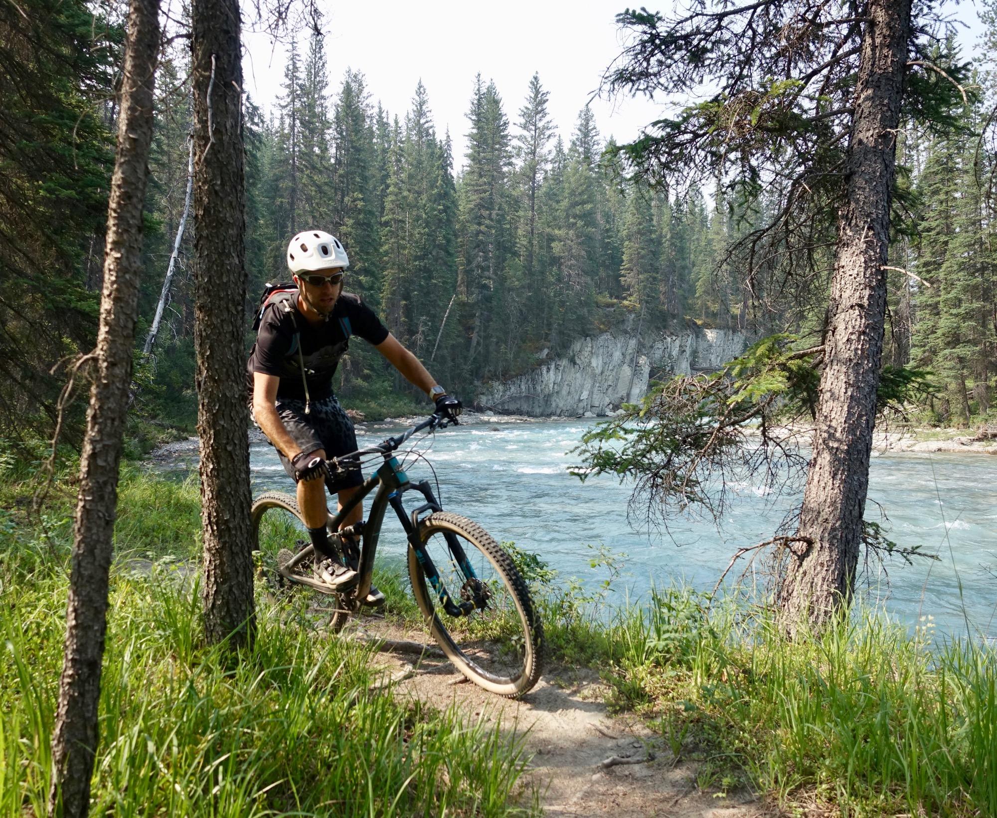 A mountain biker navigating a narrow dirt trail alongside a river, surrounded by tall coniferous trees and lush green grass. The water is a clear blue, and a rocky cliff is visible in the background. The biker is wearing a helmet and protective gear. Nipika Mountain Resort mountain bike trail.